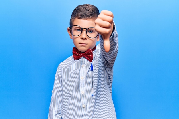 Cute blond kid wearing nerd bow tie and glasses looking unhappy and angry showing rejection and negative with thumbs down gesture. bad expression.