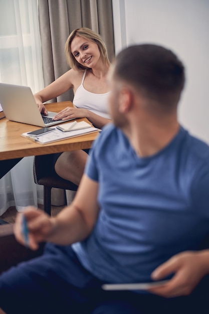 Cute blond woman smiling to brunette sporty man while sitting at table in front of laptop