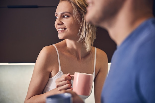 Cute blond woman smiling while staying with male in cosy atmosphere with cup of tea