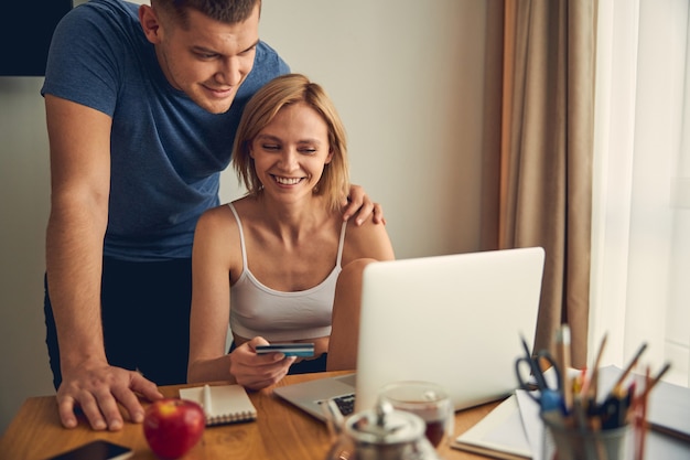 Cute blond woman working on computer from home online while brunette sporty male hugging her