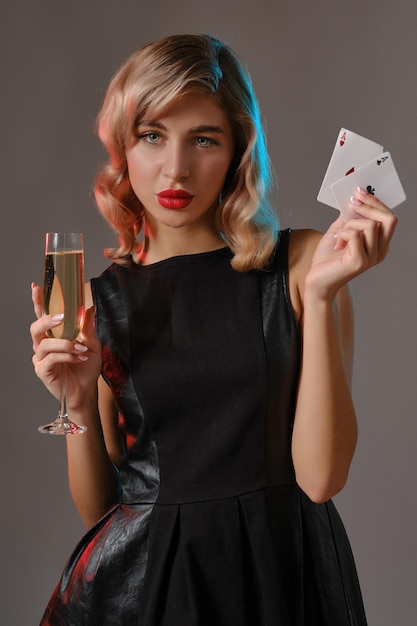 Photo cute blonde curly maiden bright makeup in black stylish dress holding two playing cards and glass of champagne posing against gray studio background gambling poker casino closeup