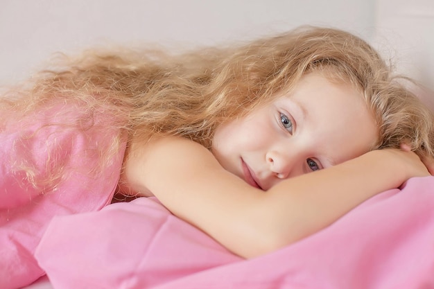 cute blonde girl with curly hair is lying on a bed with pink bed linen