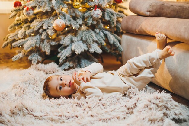 Cute boy 1 year old near the Christmas tree sitting on the floor under the tree in the room Holiday season New Year Christmas