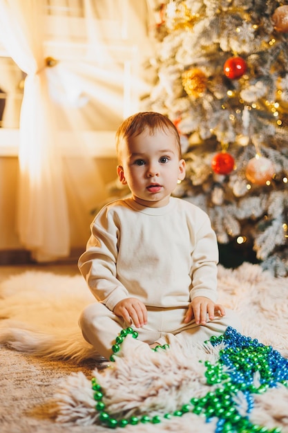 Cute boy 1 year old near the Christmas tree sitting on the floor under the tree in the room Holiday season New Year Christmas