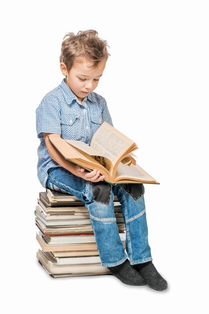 Cute boy in a blue shirt sitting on a pile of books and reading a book on a white background Isolated