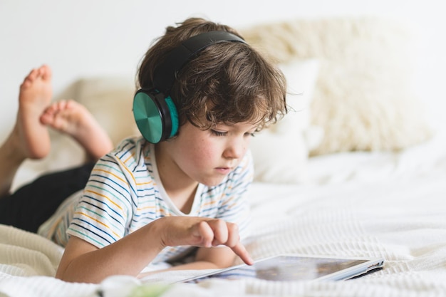 Cute boy lying on the bed at home and playing with pc tablet or reading online and listening to music with wireless headphones