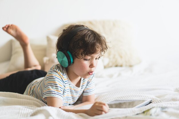 Cute boy lying on the bed at home and playing with pc tablet or reading online and listening to music with wireless headphones