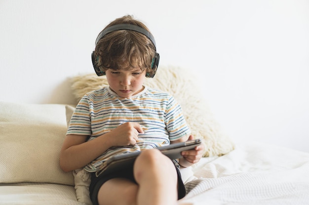 Cute boy lying on the bed at home and playing with pc tablet or reading online and listening to music with wireless headphones