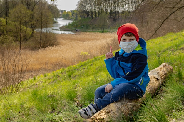 Cute boy in a medical mask sits on a hill on a log and look at the lake. Family walks with children outdoors in early spring during quarantine