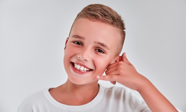 Cute boy put his hand to his ear saying call me on a gray background and smiling.