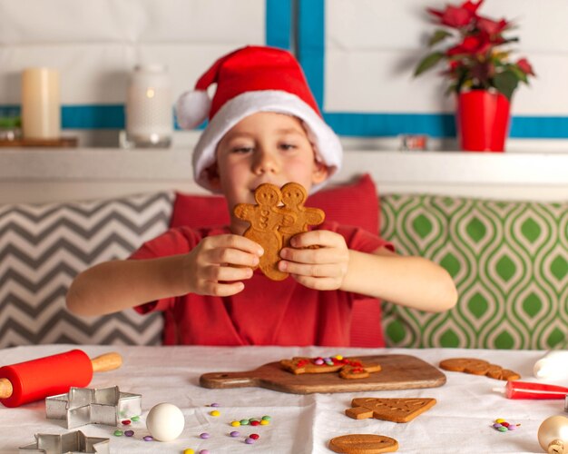 Cute boy in santa claus hat makes gingerbread at the table christmas lights