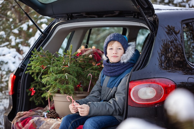 Cute boy sitting in black car at snowly winter forest. Christmas concept.