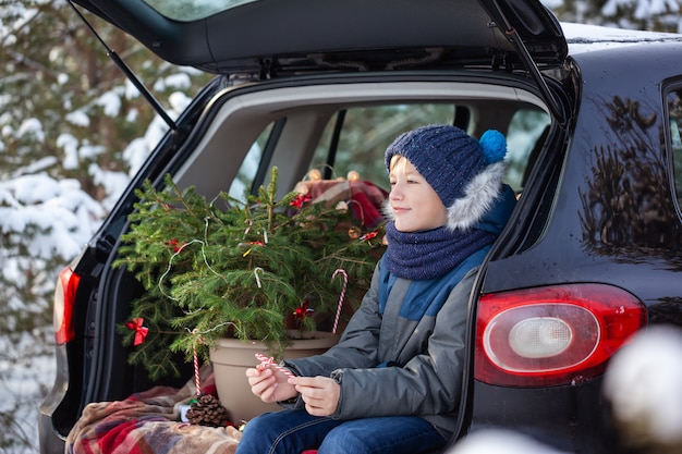 Cute boy sitting in black car at snowly winter forest. Christmas concept.