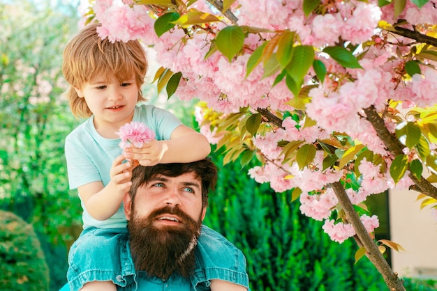 Cute boy sitting on fathers shoulders father giving son ride on back in park happy childhood concept