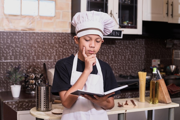 Cute boy using chef uniform is thingking while reading a book recipe prepare for cooking in the kitc