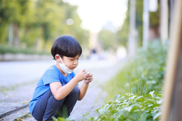 A cute boy wearing a blue shirt long black pants and a white mask sits in a lush green garden in the spring against a backdrop of green trees portrait shot