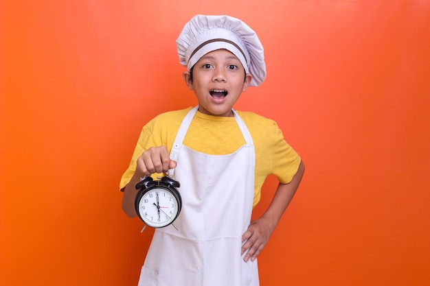 Cute boy wearing chef uniform gesturing shocked expression while holding alarm clock