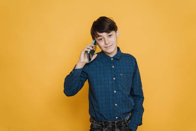 Cute boy with brown eyes dressed in casual shirt and jeans making a call with a new cellphone, looking confident and satisfied against yellow backdrop with a copy space. Comunication people concept.