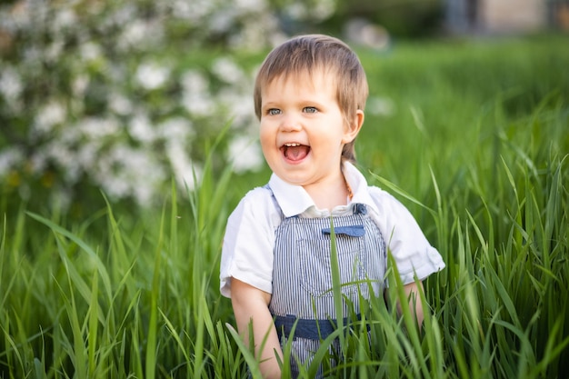 A cute boy with expressive blue eyes in a fashionable jumpsuit smiles funny and hides in the tall green grass