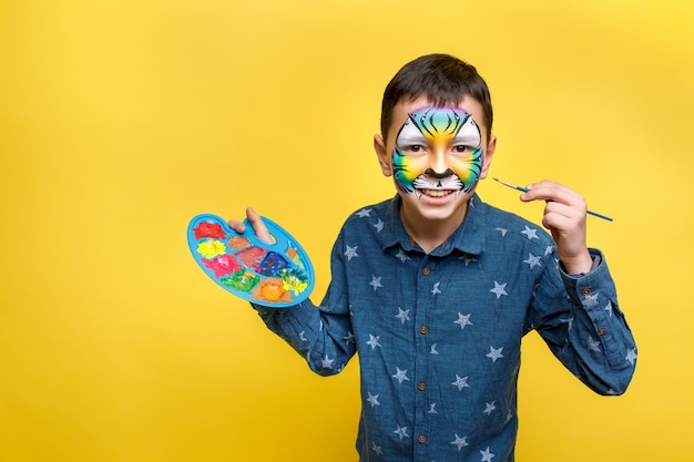 Cute boy with faceart on birthday party, colorful tiger holding palette with gouache isolated on yellow wall.