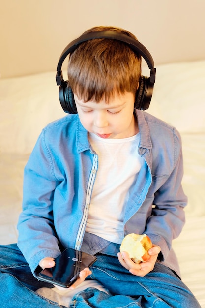 Cute boy with phone and head phones listening music indoors sitting on the bed in the bedroom Child eats an apple