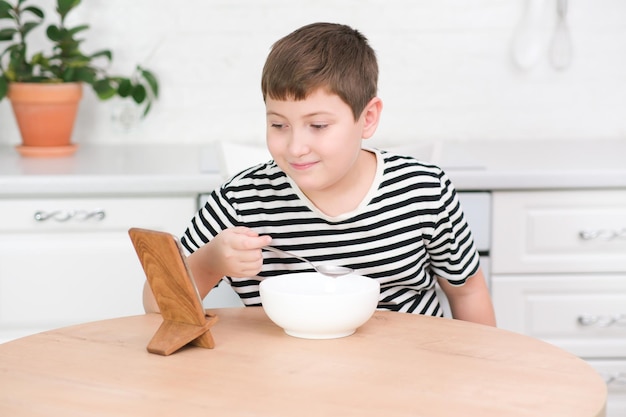 A cute boy of years old watch mobile phone cartoons and eat at round wooden table in light kitchen