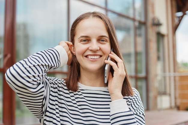 Cute brown haired looking at camera while talking on smartphone wearing striped shirt holding cell phone pleasant conversation while resting outdoor