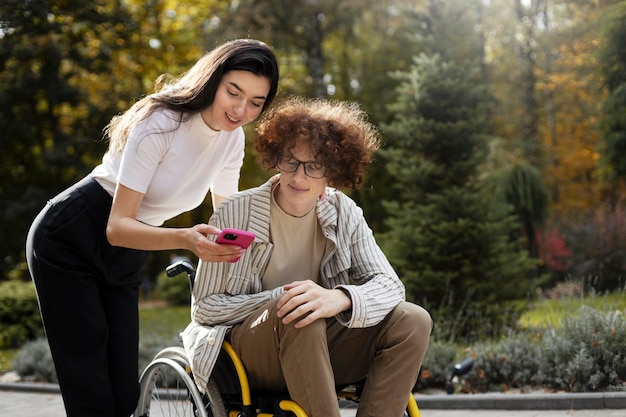 Cute brunette girl shows her phone to her disabled friend A young man sits in a wheelchair outdoor