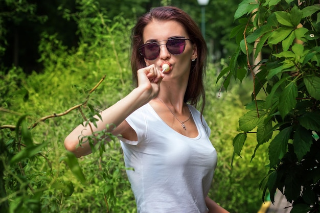 Cute brunette in glasses stands near a tree and licks a lollipop