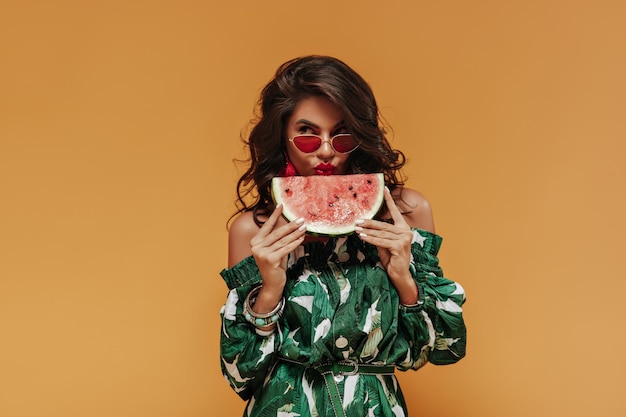 Cute brunette haired lady with bright lips in unusual accessories and fashionable green dress posing with watermelon and looking away