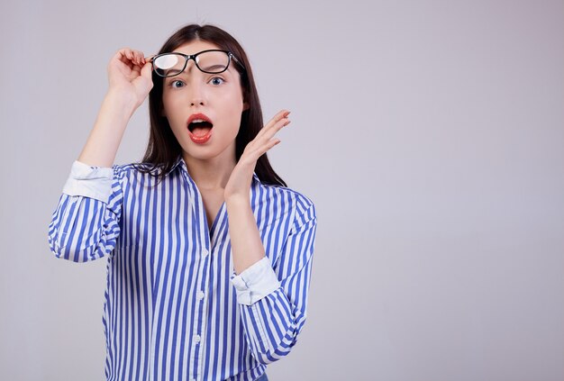 Photo cute business woman with brown hair, full pink lips posing. she is wearing a white and blue striped shirt and black computer glasses. surprised. looks sideway. copy space.