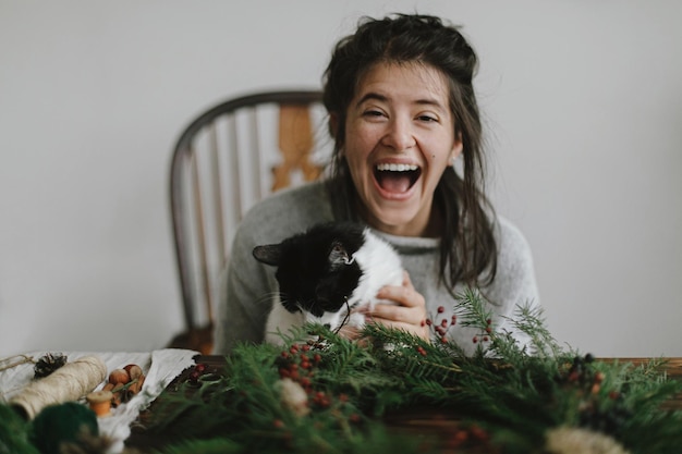 Cute cat helping young happy woman making rustic christmas wreath Funny authentic home moments