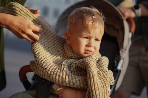 Cute caucasian baby boy sitting in a stroller. mother is putting a swaeter on him.Image with selective focus. High quality photo