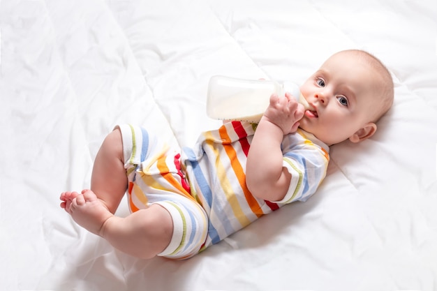 Cute caucasian baby with first baby milk bottle. Top view. Artificial feeding concept. Baby milk formula. Infant in striped jumpsuit on white blanket.