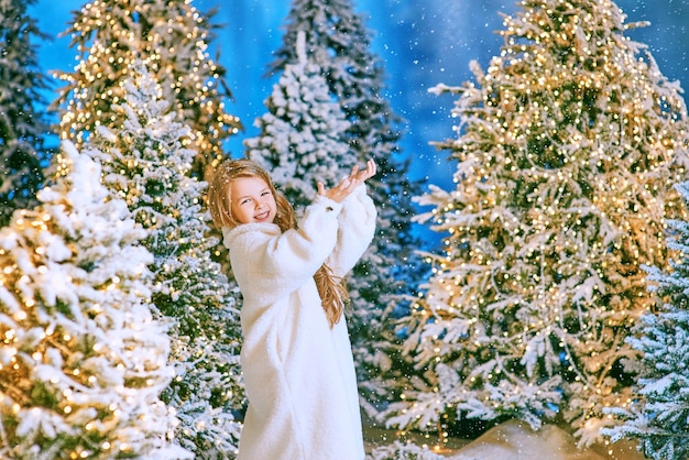 cute caucasian blonde girl in white eco fur coat walking in winter christmas forest with lights.