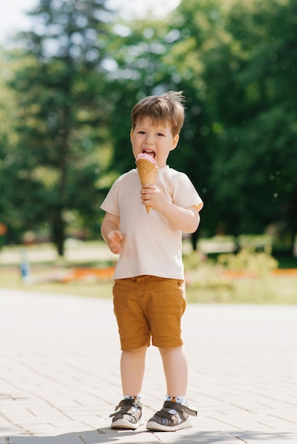 Cute Caucasian boy of three years old eats ice cream in a cone while walking in a summer park