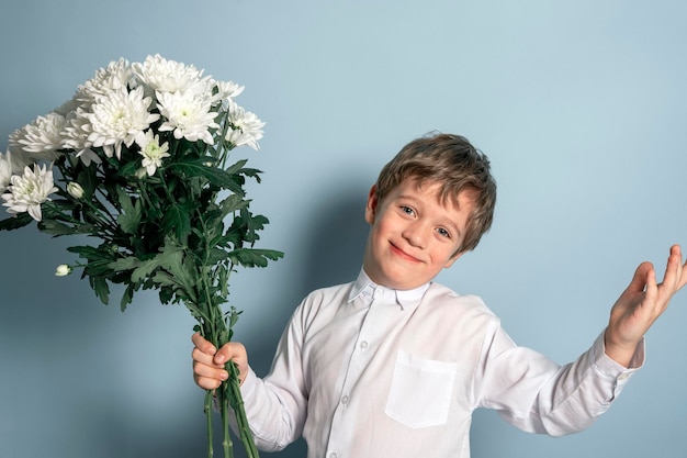 A cute Caucasian boy in a white shirt holds a bouquet of white flowers in his hand and smiles