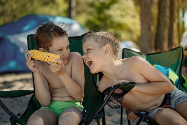 Cute caucasian boys eating corn on beach sitting on camping chairs laughing enjoyig holiday