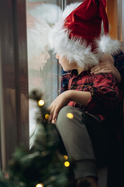 Cute caucasian children wearing snta hats and plaid shirts sitting hugging by the window waiting for christmas. christmas tree with lights bokeh on foreground. Reflection in window