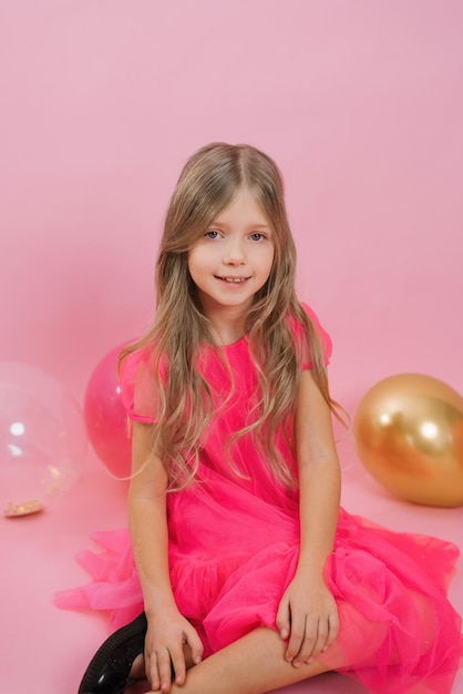 Cute Caucasian girl of seven years old in a bright pink barbicore style dress sits on a pink background with festive water balloons for her birthday