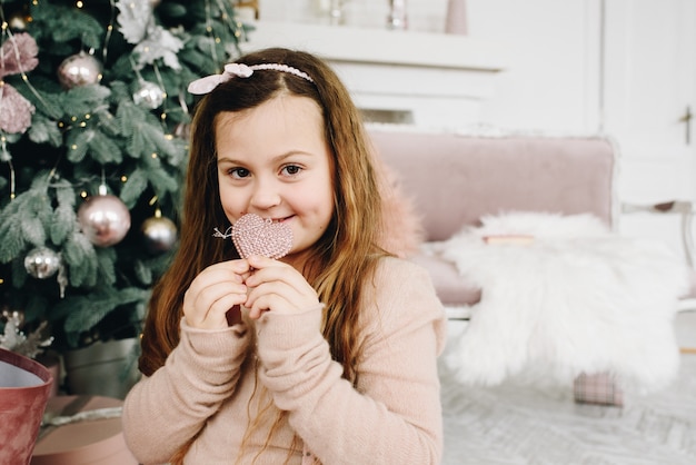 Cute Caucasian girl sitting near Christmas Tree holding a heart-shaped decoration