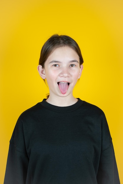 Cute Caucasian kid girl wearing black sweatshirt against yellow wall showing tongue