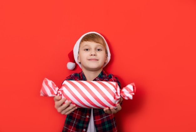 Cute caucasian kid teenager boy in Santa hat holds candy shape gift box with surprise on red background Copy space for design or text Banner Christmas New Year mockup template