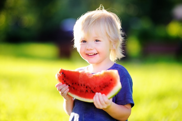 Cute caucasian little boy with blond hairs eating fresh watermelon outdoors