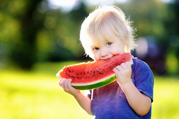 Cute caucasian little boy with blond hairs eating fresh watermelon outdoors