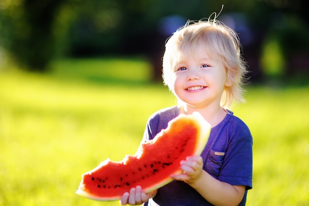 Cute caucasian little boy with blond hairs eating fresh watermelon outdoors