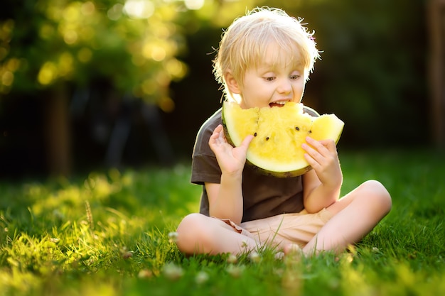 Cute caucasian little boy with blond hairs eating yellow watermelon outdoors