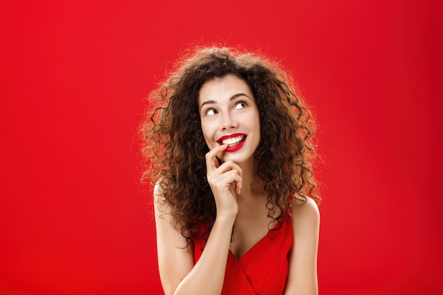 Cute charismatic and tender dreamy woman with curly hairstyle in red dress biting finger and smiling...