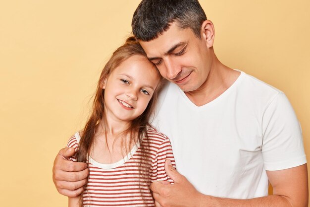 Cute charming gently family father and daughter wearing casual tshirts standing isolated over beige background dad hugging kid with love closed eyes