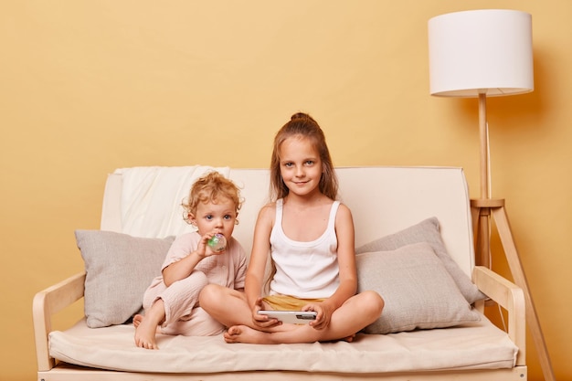 Cute charming little girls sisters sitting on couch using smartphone together posing against beige wall playing online games spending free time with gadget